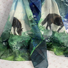 A lightweight sheer scarf draped over a patterned white chair, featuring a watercolor-style print of two dark horses in a green forest landscape with blue sky and white blossoms.