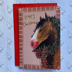 A Christmas greeting card featuring a brown horse with a green wreath and red bow, layered over a red envelope, placed on light blue wrapping paper with a repeating horse pattern.
