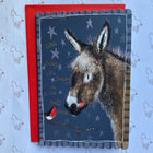 A square greeting card featuring an illustrated donkey in profile against a dark blue starry background, with a small robin and gold text reading 'Little Donkey, Little Donkey on the daily road.' The card is layered over a red envelope and placed on a light blue patterned surface.