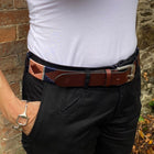 A person wearing a fitted white short-sleeve scoop-neck t-shirt, black shorts, and a brown leather belt with a colorful patterned section, standing outdoors against a brick and stone wall.