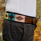 A close-up of a person wearing a brown leather belt with a colorful geometric patterned keeper and black pants, standing outdoors against a stone wall and fallen leaves.