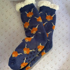 A pair of navy blue knit socks with orange and cream fluffy highland cows featuring grip dots on the soles and a cream sherpa trim at the top, displayed on a patterned white background.