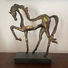 A bronze-colored abstract horse sculpture with a gold-painted back, standing on a dark rectangular base on a wooden table against a white wall.