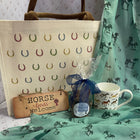 A horse-themed gift set featuring a cream canvas tote bag with colorful horseshoe patterns, a mint green scarf with horse silhouettes, a wooden sign reading 'Horse Lovers Welcome', a small wrapped gift, and a white ceramic mug with horse illustrations.
