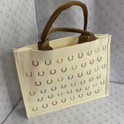 An off-white canvas tote bag with brown handles, decorated with a grid of small colorful horseshoes in green, blue, and red, set against a patterned backdrop.
