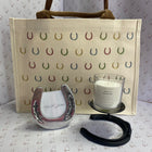 A horse-themed gift set featuring a canvas tote bag with colorful horseshoe patterns, a polished silver gallopers horseshoe, a black leather bridle bit, and a white scented candle in a glass jar, all arranged on a patterned white surface.