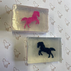 2 bars of clear soap containing a pink horse and a black horse toy on a grey background