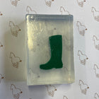 A clear rectangular soap containing a dark green boot-shaped object, placed on white paper printed with a repeating brown horse outline pattern.