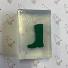 A small clear rectangular soap containing a dark green boot-shaped object, placed on white paper printed with a repeating brown horse outline pattern.