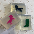 Three translucent rectangular soap bars with horse-themed designs, including a black horse, a pink horse, and a green boot, arranged on a white background with a repeating horse outline pattern.