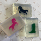 Three translucent rectangular soap bars with horse-themed designs, including a black horse, a pink horse, and a green boot, arranged on a white background with a repeating horse outline pattern.