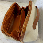 An open canvas cosmetic bag with a cream-colored woven exterior, tan leather trim, and a warm brown interior, shown on patterned paper.