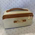 A structured canvas cosmetic bag with a cream-colored woven body, tan leather handle and trim, and gold-tone hardware, including a horsebit detail on the front and zipper pulls. 