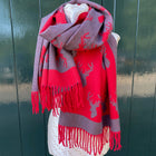 A red and gray knit scarf with deer silhouettes and fringe, draped on a white dress form against a dark green wooden fence.