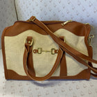 A brown and beige canvas duffel bag with gold-tone hardware, featuring a horsebit detail on the front and a shoulder strap, photographed against a patterned backdrop.