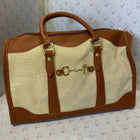 A structured saddle-style handbag with tan leather trim and handles, a beige canvas body, and gold-tone horsebit hardware, photographed against a patterned backdrop.