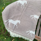 A taupe wool blanket with white horse motifs and white fringe, draped over a wooden bench on a grassy lawn.