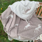 A woven blanket with white horse motifs and white fringe, draped over a wooden chair on grass.