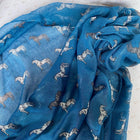 A blue sheer fabric scarf or shawl draped in soft folds, printed with a repeating pattern of gray and white horses, laid flat on a light patterned surface.