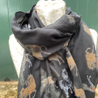 A dark charcoal gray scarf with a pattern of horses in tan, white, and gray, draped on a cream-colored mannequin torso against a green wooden background.