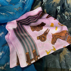 A folded pink silk scarf with a rustic equestrian theme, featuring brown horses, wooden fences, and striped bands, displayed on top of other patterned scarves.