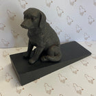 A dark gray dog figurine sitting on a rectangular black base, photographed against a white backdrop with a repeating horse outline pattern.