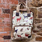Pale green and brown rucksack with handles and a front zip pocket. It has red foxes, black Labradors, pheasants, deer and rabbits on it. It is hung on a brick wall. - Gallop Guru