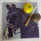 A square gray serving board featuring a horse head mosaic made from purple grapes, beads, and white grapes, styled with a green apple, a knife, and a avocado on top, set against a white patterned background.