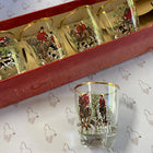 A vintage set of five clear glass shot glasses with gold rims and a hunting scene printed on them, shown in an open red box with one glass placed in front on patterned paper.