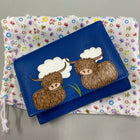 Bright blue folded purse with two highland cows sat beneath the clouds eating grass appliqued on the front. Shown on a coloured flower pouch that the purse comes in.